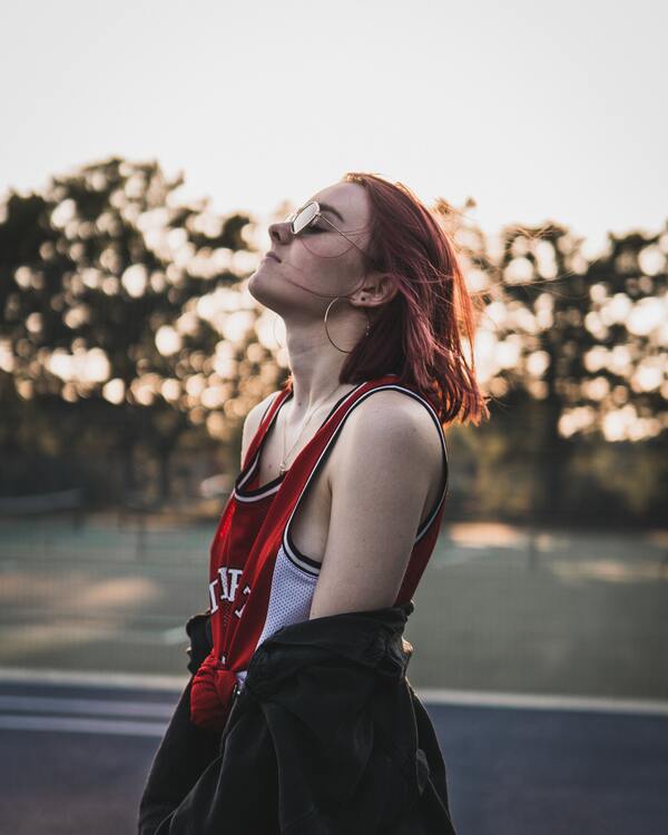 Mulher branca e ruiva, posando de lado com olhos fechados e óculos escuros. Ela veste uma regata de basquete. 